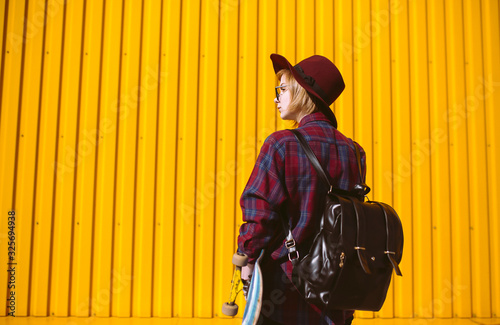 Young teenager girl with skateboard in hand posing on a yellow background. Pretty woman in sunglasses and hat stands on the street with skateboard in hand. Lifestyle, youth concept.