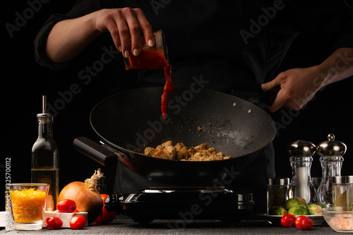 Chef adds tomato sauce to a wok with meat and vegetables.Freezing in motion. Restaurant menu, cooking.On a black background, photo for advertising or design, restaurant business, recipe book