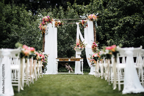 Decorated luxury wedding ceremony place in the garden. White empty chairs and arch decorated with flowers.