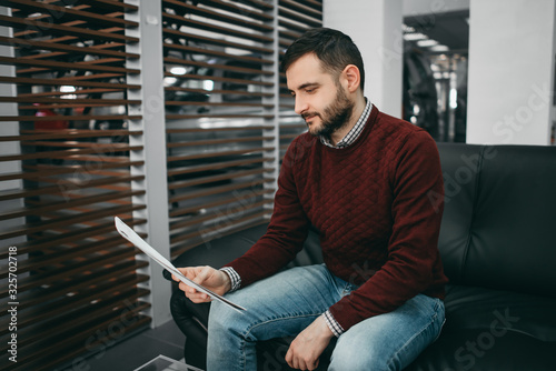 A brunette bearded man sits on a couch and reads a contract in a saloon. Happy, worried, skeptic, car loan, car rental, car purchase in salon