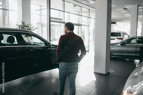 A white bearded brunette man walks in a car shop, salon, chooses a car to buy
