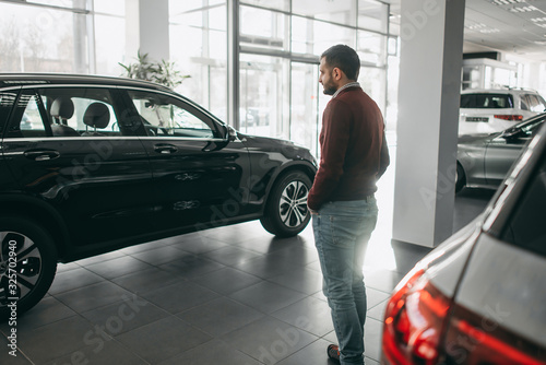 A white bearded brunette man walks in a car shop, salon, chooses a car to buy