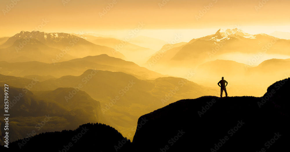 Spectacular mountain ranges silhouettes. Man reaching summit enjoying freedom. Sunrise with orange light.