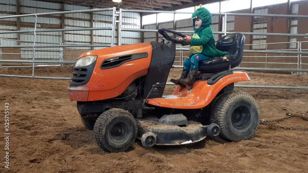 Two Year Old Toddler in Dino Costume and Cowboy Boots Sitting Playing on Riding Lawn Mower Inside Arena. Two Year Old Toddler in Dino Costume and Cowboy Boots Sitting Playing on Riding Lawn Mower Inside Arena.