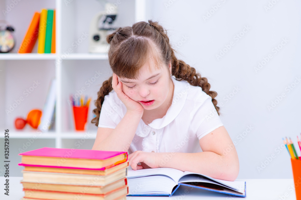 A girl with down syndrome sitting at a table at home reading a book lying in front of her A girl with down syndrome sitting at a table at home reading a book lying in front of her