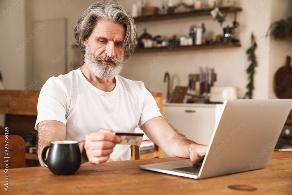 Image of serious mature man working with laptop and holding credit card