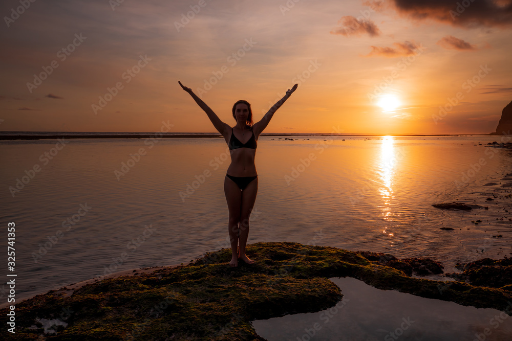 Excited young woman raising arms at the beach in front of the ocean. Sunset golden hour at the beach. Bali, Indonesia.