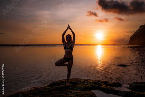 Vrikshasana asana. Young woman practicing tree pose at the beach during sunset. Arms raising with namaste mudra. Melasti beach, Bali.