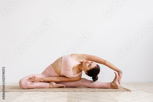 Beautiful well-groomed young woman fitness instructor in a pink suit doing a warm-up stretching while sitting on a rug on the floor barefoot on a white background. Place for advertising