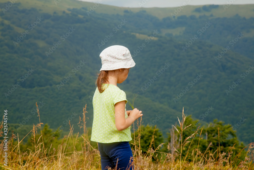 Portrait of cute little child girl in the grass against mountain background. Happy child have a rest in mountains.