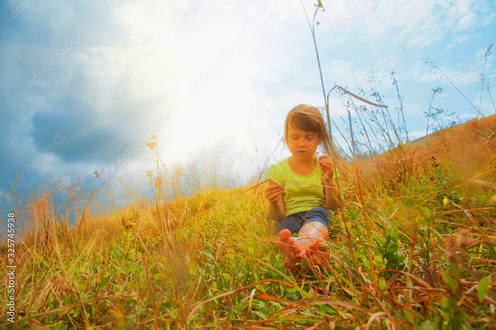 Cute little child girl sitting in dry grass against mountains under sunlight on sunny day.  Happiness,  childhood and  carefree concept.