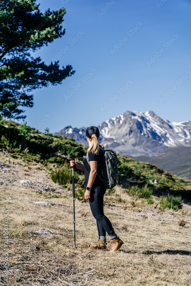 A blonde woman with a cap and backpack, trekking through a landscape of meadow and trees with snowy mountains in the background
