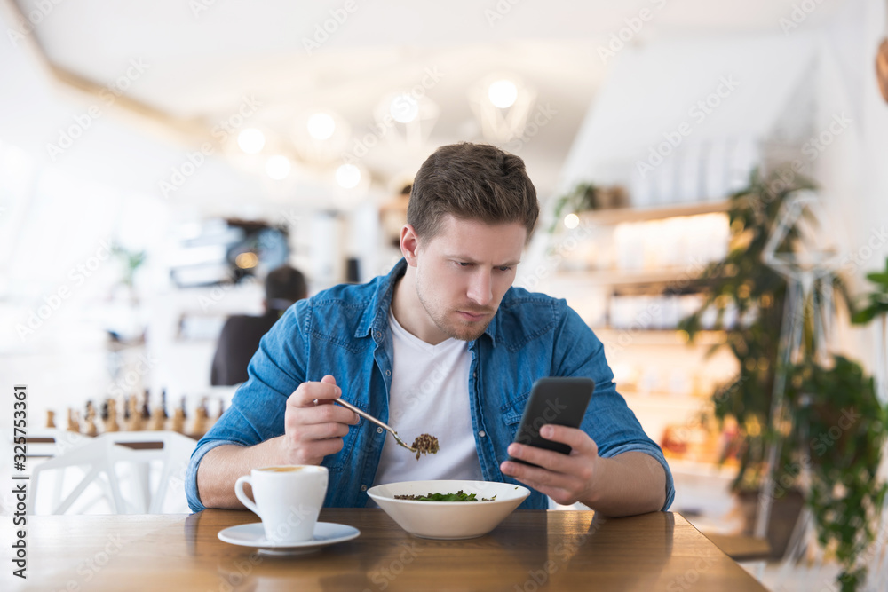 Young handsome smiling man drinks his hot coffee and eats salad for lunch while typing message in his smartphone during break at cafe , multitasking concept Young handsome smiling man drinks his hot coffee and eats salad for lunch while typing message in his smartphone during break at cafe , multitasking concept
