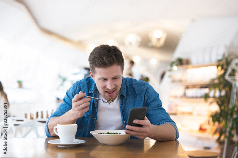 Young handsome smiling man drinks his hot coffee and eats salad for lunch while reading message in his smartphone during break at cafe , multitasking concept