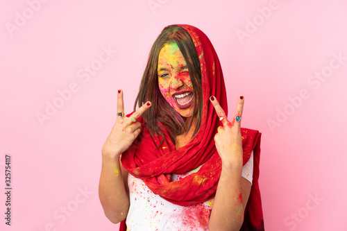 Young Indian woman with colorful holi powders on her face isolated on pink background smiling and showing victory sign