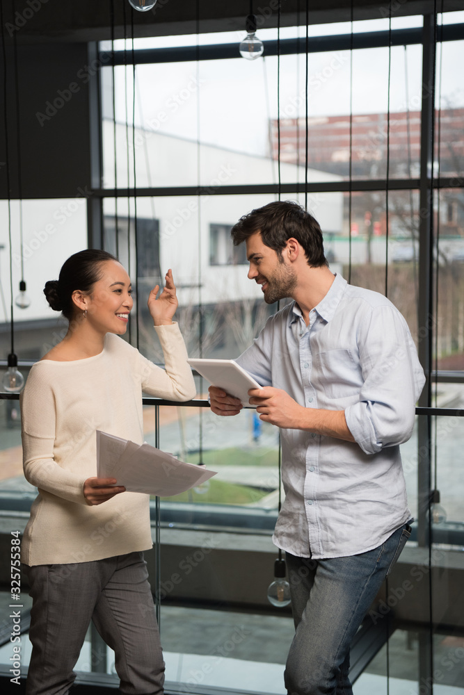 Obraz premium cheerful asian businesswoman holding papers and talking to handsome colleague with digital tablet