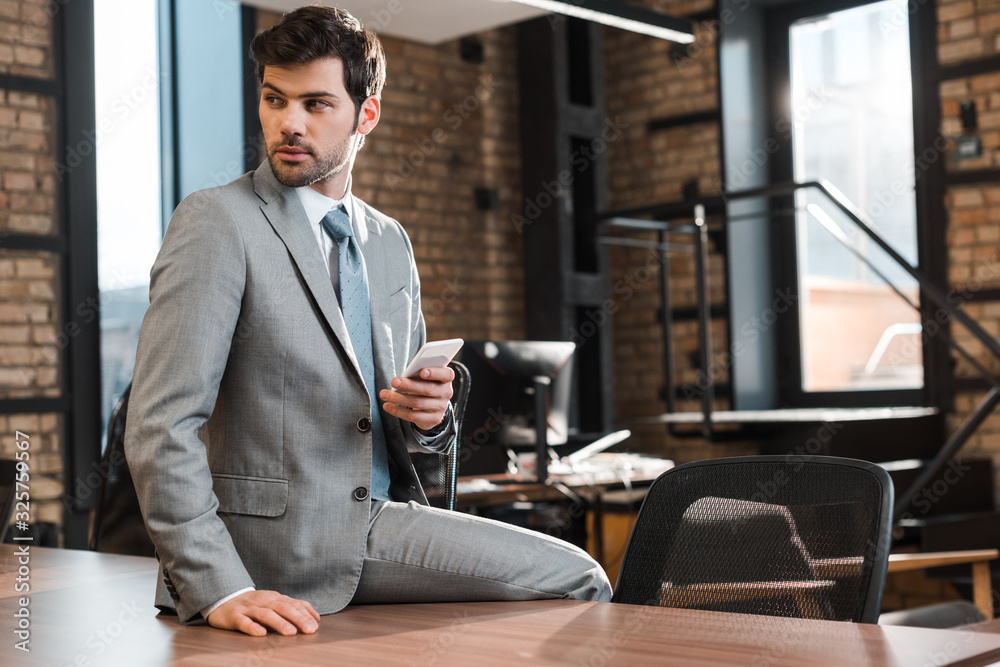confident, handsome businessman sitting on office desk, holding smartphone and looking away confident, handsome businessman sitting on office desk, holding smartphone and looking away