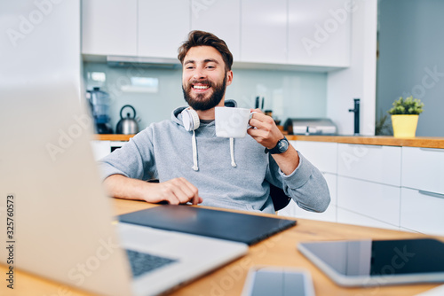 Joyful young web developer drinking coffee while taking break from work in home office