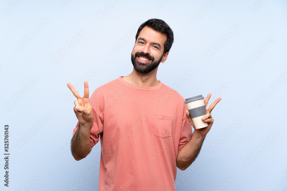 Young man with beard holding a take away coffee over isolated blue background showing victory sign with both hands Young man with beard holding a take away coffee over isolated blue background showing victory sign with both hands