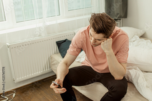 tired young caucasian guy at morning after party night, man sit on bed at home and look at smartphone. isolated in bedroom © Roman