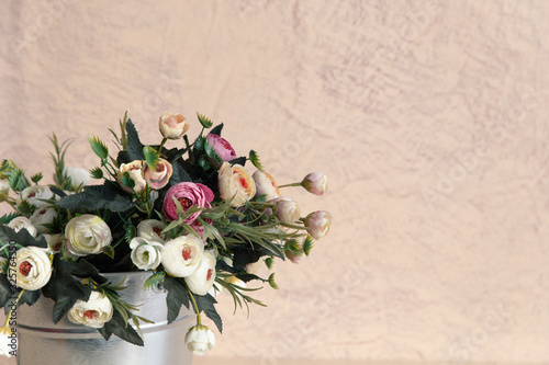 Beautiful white, beige and pink flowers in close-up stand in a decorative aluminum bucket against the beige wall in the left corner. Space for text, postcards. Concept of a postcard.