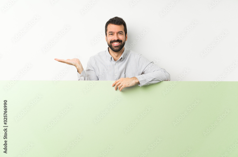 Young handsome man with beard holding a big green empty placard holding copyspace imaginary on the palm to insert an ad Young handsome man with beard holding a big green empty placard holding copyspace imaginary on the palm to insert an ad