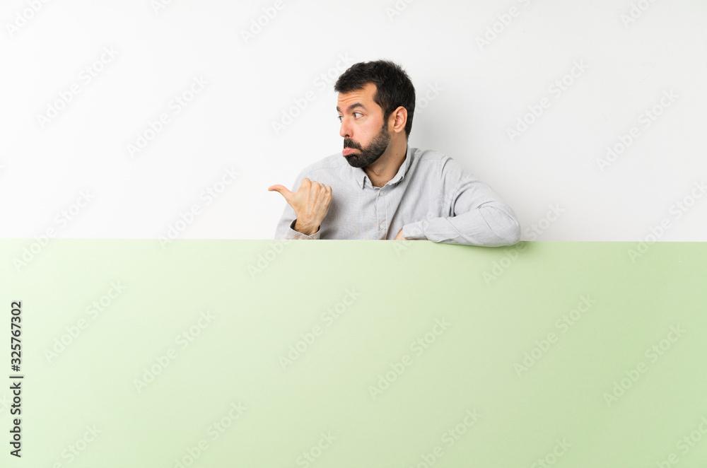 Young handsome man with beard holding a big green empty placard unhappy and pointing to the side Young handsome man with beard holding a big green empty placard unhappy and pointing to the side