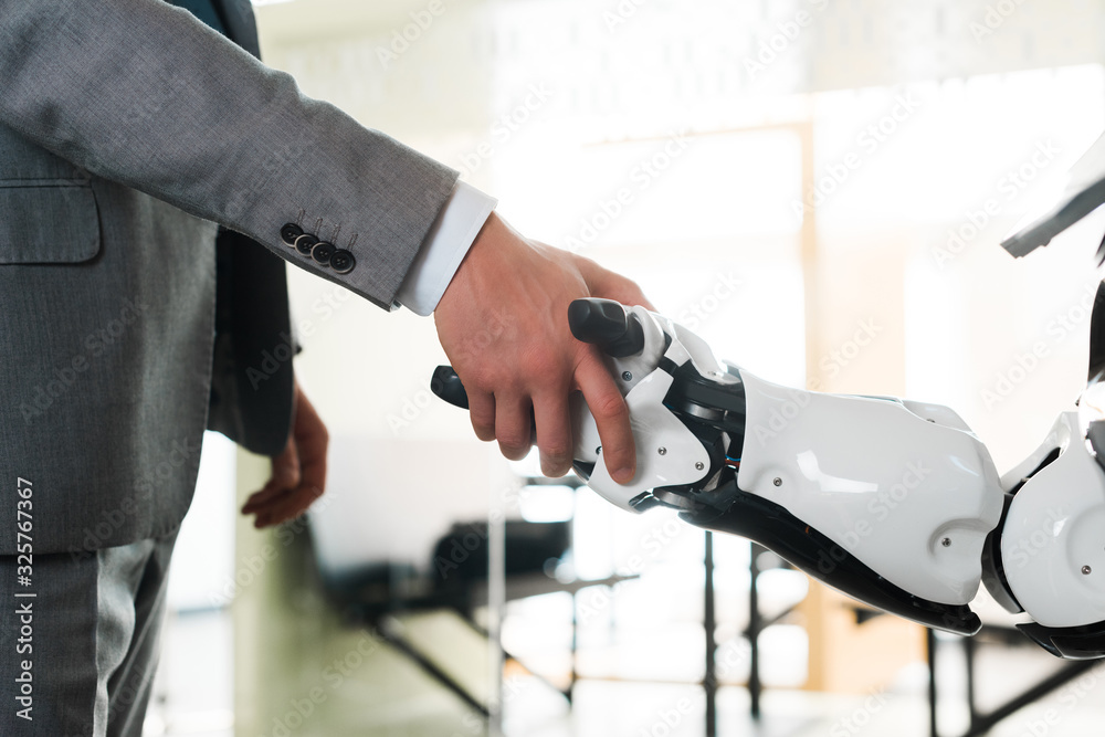 cropped view of businessman and robot shaking hands in office cropped view of businessman and robot shaking hands in office