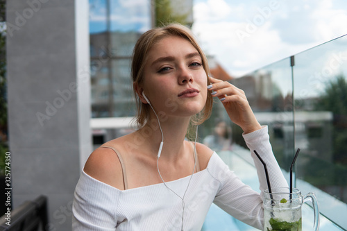 Porteait of blond woman blogger with cocktail is listening music or e-book on smartphone connected to free wifi in cafe. Positive young student listening podcasting online on modern mobile phone. photo