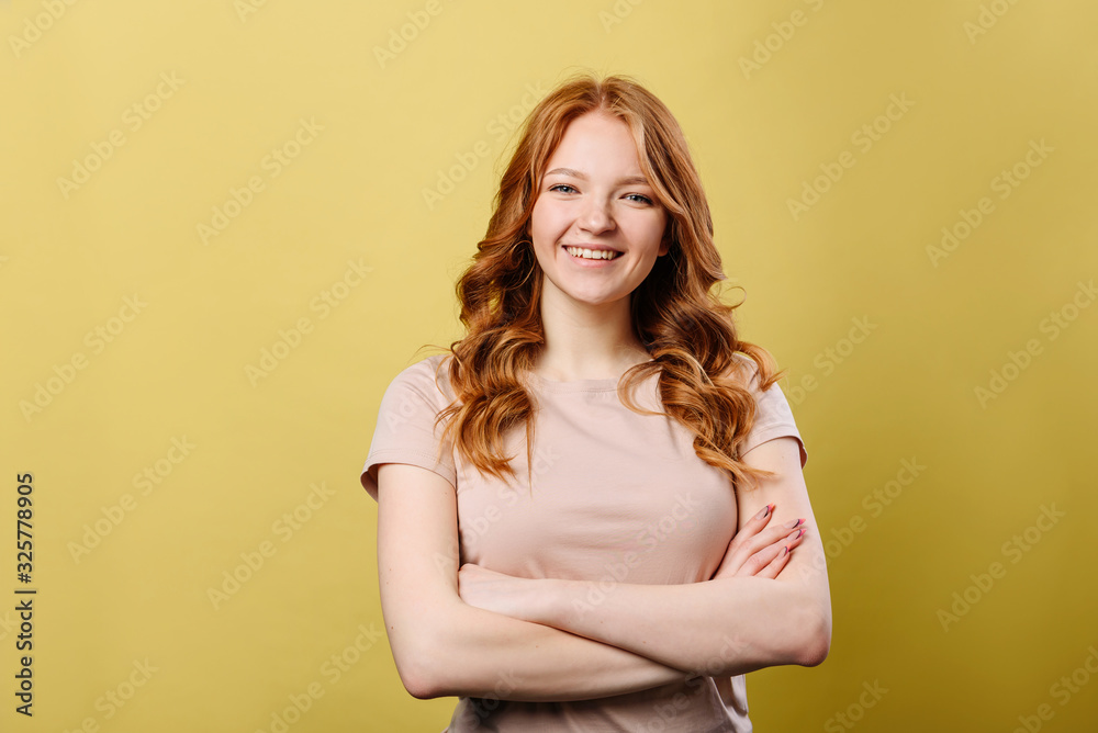 Image of a beautiful, red-haired, smiling, emotional girl posing with arms folded across her chest. A girl is standing on a yellow background. Image of a beautiful, red-haired, smiling, emotional girl posing with arms folded across her chest. A girl is standing on a yellow background.