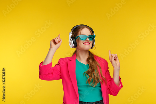 Enjoy listening to music. Young redhead woman in headphones listening music. Funny smiling girl in earphones and pink jacket dancing and singing on yellow background. Relaxation and stress management.