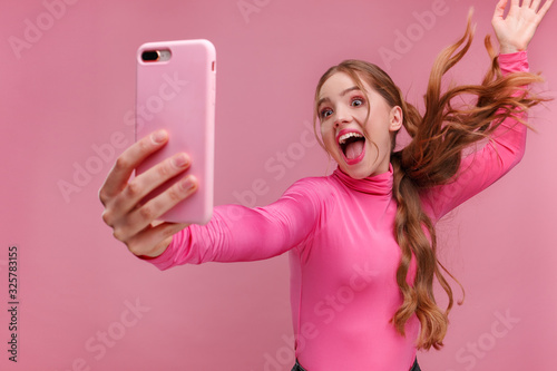 Funny young redhead woman making selfie. Smiling girl wearing pink blouse holding pink smartphone, making faces on camera, posing for selfie isolated on pink background. photo