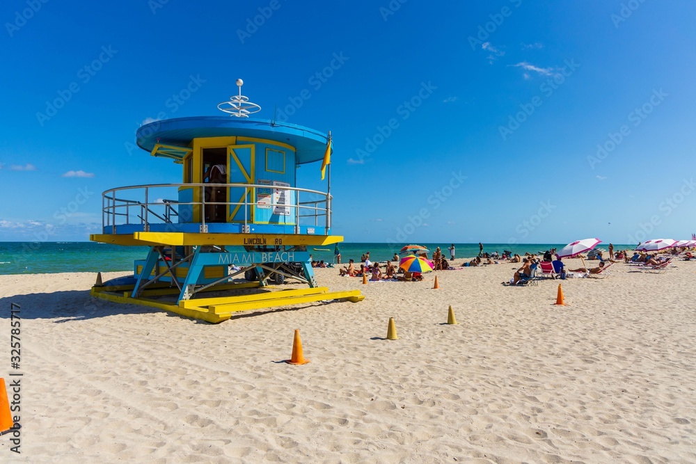 People on Miami beach on beautiful sunny day. Sand beach, tourists and yellow lifeguard tower on blue Atlantic ocean merging with blue sky background. Miami. USA.