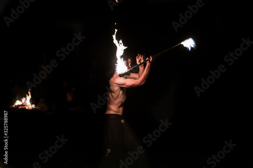A side profile view with selective focus on a muscular fire dancer performing with a flaming stick outdoors by night, details of the arm and torso muscles working