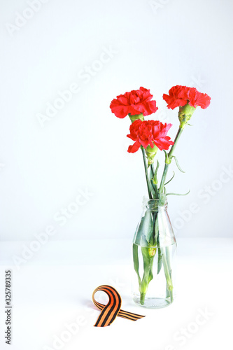 Three red carnations and St. George ribbon (Russian symbol) on a white uniform background. Greeting card for the holiday of the great victory of May 9.
