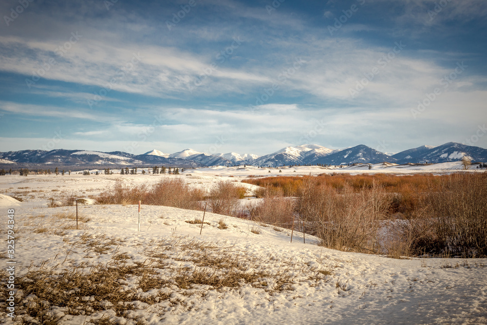 Vast mountain range behind wide open snow covered field with brush and vintage fencing on clear day
