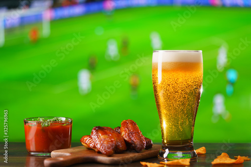 Spicy barbecue chicken wings, tomato salsa, nachos and beer on dark wooden bar table. Football on background, high resolution