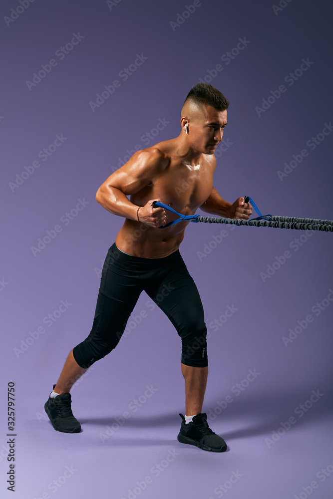 strong shirtless man training with expander, guy is keen of fitness, man preparing for a competition, full length side view photo. isolated blue background, studio shot strong shirtless man training with expander, guy is keen of fitness, man preparing for a competition, full length side view photo. isolated blue background, studio shot