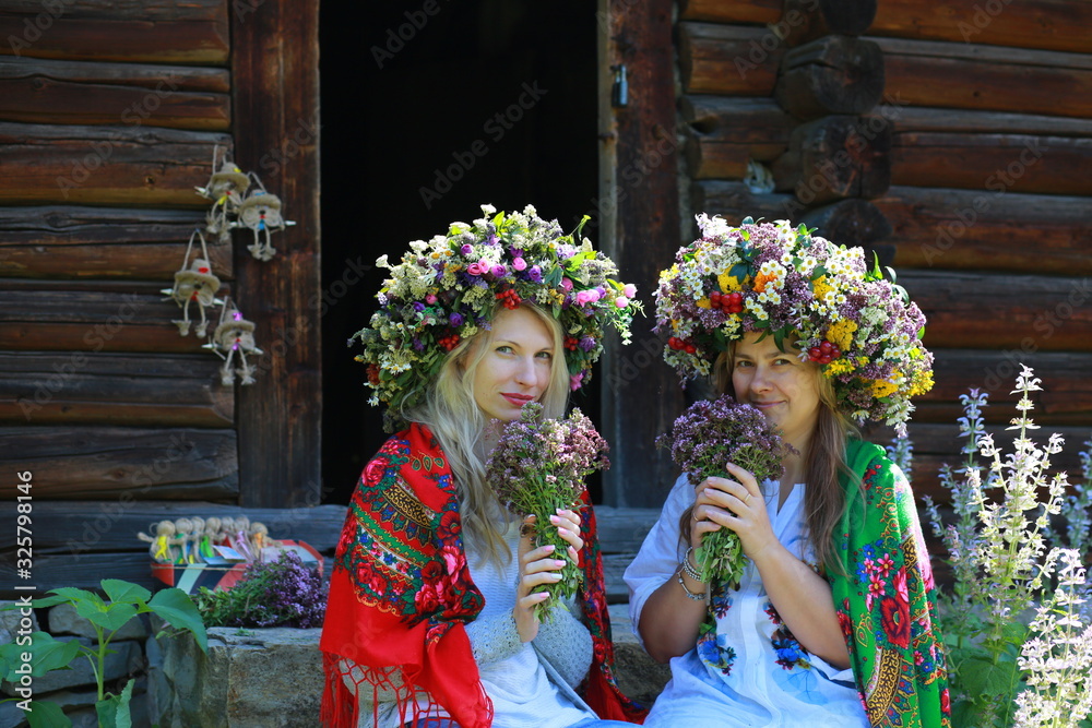  two girl friends in floral wreaths and scarves in Ukrainian ethnic style enjoy the aroma of flowers