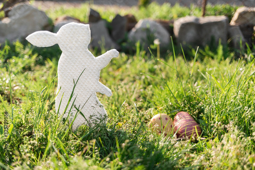 The concept of Easter holidays. Decorative gift handmade product in the form of a rabbit, located next to the painted eggs. Green grass and stones in the background. Copy space