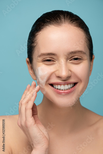 smiling girl applying cleansing foam on face, isolated on blue