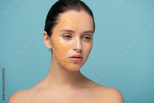 attractive girl applying yellow sugar exfoliant on face, isolated on blue