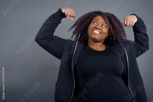Waist up shot of African woman raises arms to show her muscles feels confident in victory, looks strong and independent, smiles positively at camera, stands against gray background. Sport concept.