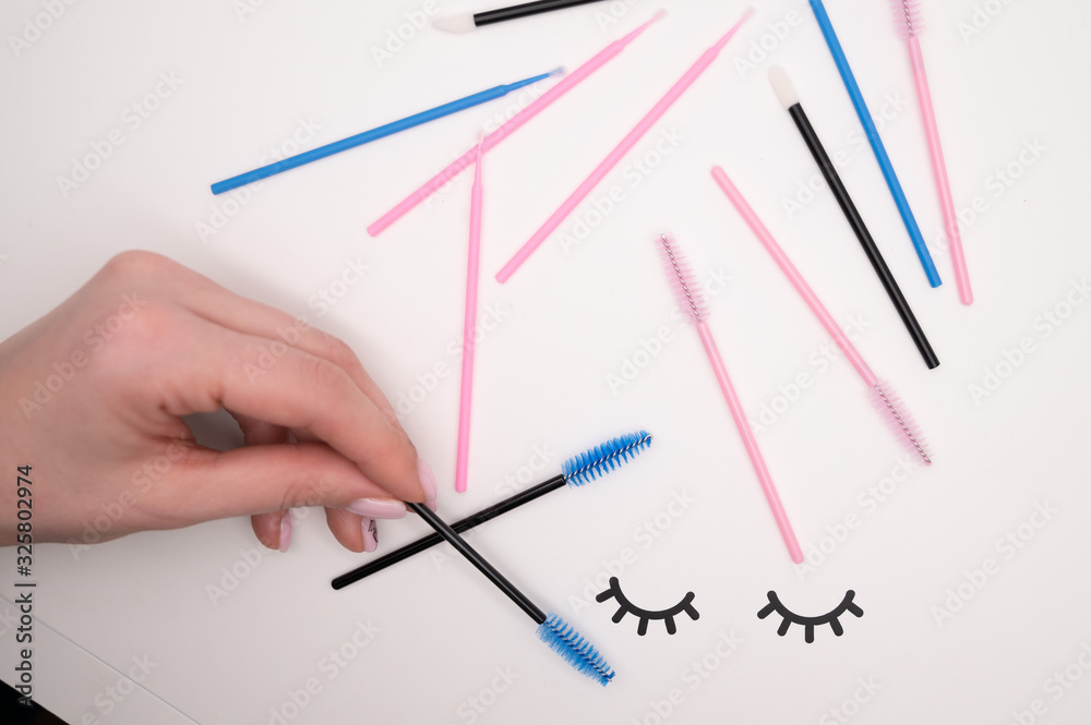 Top view of flatlay from eyelash extension tools. Female hand holds colored brushes for combing cilia.
