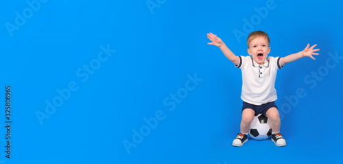 Boy sits on soccer ball and rejoices in victory, raising his arms to top against blue background. Emotions of winner and champion, place for text. Concept of winning and achieving goal.