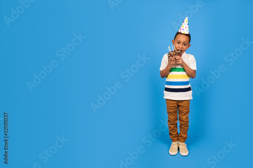 Happy African-American boy with a funny little party cone on his head celebrates birthday holding chocolate cake in his hands and biting cake rejoicing in a blue background. Birthday party concept