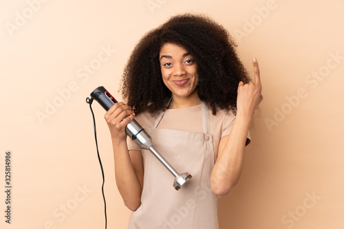 Young african american woman using hand blender isolated on beige background pointing with the index finger a great idea