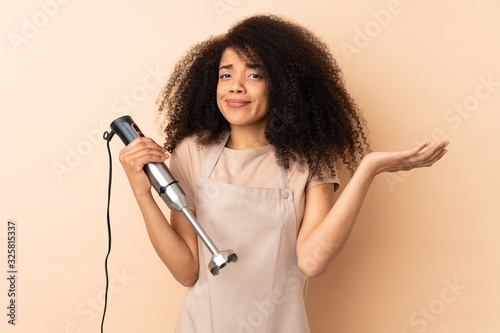 Young african american woman using hand blender isolated on beige background having doubts with confuse face expression