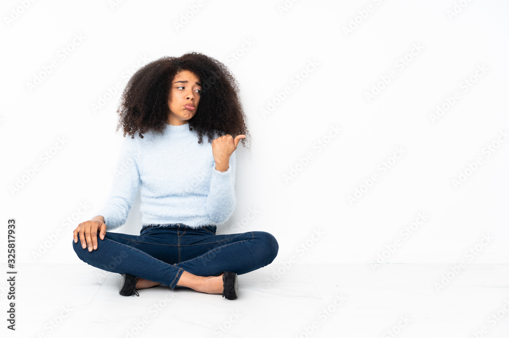 Young african american woman sitting on the floor unhappy and pointing to the side Young african american woman sitting on the floor unhappy and pointing to the side