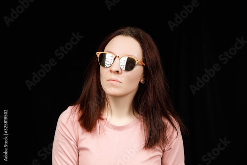 Image of sweet confident young lady standing over black background in studio, wearing pink blouse and fashionable eyeglasses, having long brown hair